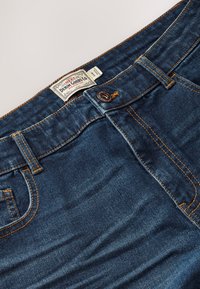 Dark blue denim jeans with orange stitching, a button closure, belt loops, and a fabric label reading "Denim Goods Co." near the waistband.