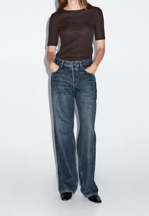 Woman wearing a fitted dark brown short-sleeve top, high-waisted wide-leg blue jeans, and black pointed shoes, hands in pockets.