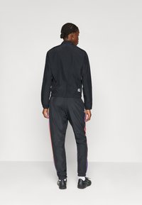 Μαύρο μπουφάν αδιάβροχο με ελαστικές μανσέτες και στρίφωμα, συνδυασμένο με μαύρο παντελόνι φόρμας που έχει χρωματιστές ρίγες στο πλάι.