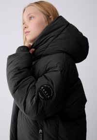 Zwarte pufferjas met een hoge capuchon, gestructureerde stof en elastische manchetten. Heeft een ronde logo-patch op de mouw en een ritssluiting.