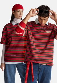 Camisetas gigantes a rayas en rojo, negro y amarillo, con logos. El modelo masculino lleva gafas de sol; la modelo femenina lleva una gorra roja.