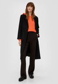Zwarte lange jas over een oranje hoodie, gecombineerd met zwarte broek en chunky zwarte schoenen. De jas heeft een notch-kraag en zakken.