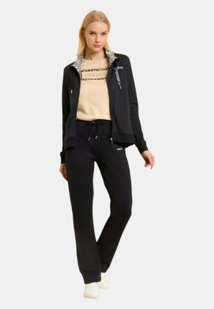 Donna che indossa una camicia beige e una giacca nera con zip e colletto stampato leopardato, pantaloni neri con coulisse e sneakers bianche, in piedi su sfondo bianco.