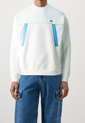 Personne portant un sweat-shirt blanc avec des accents bleus et verts et un jean cargo bleu, debout devant un fond clair uni.