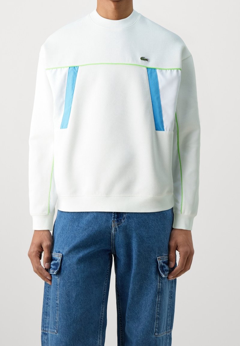 Personne portant un sweat-shirt blanc avec des accents bleus et verts et un jean cargo bleu, debout devant un fond clair uni.
