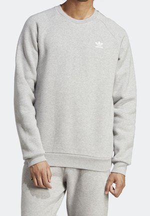 Man draagt een lichtgrijze Adidas trui en bijpassende joggingbroek met kleine witte Adidas-logo's op borst en dij.