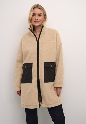 Beige fleece coat with a zip front, high collar, and two black fabric pockets. Features a soft texture and a relaxed fit.