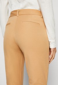 Lichtbruine broek van een gladde stof, met een taillering in de taille en twee achterzakken, gecombineerd met een wit longsleeve shirt.