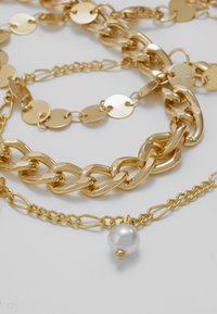 Auksinės grandinėlės su įvairiais ryšiais, blizgiu paviršiumi ir viena balta perlo akcentu ant subtilios grandinėlės.