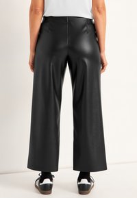 Brede zwarte faux leren broek met een gladde textuur, voorzien van een hoge taille en subtiele gemiddelde naden. Gecombineerd met zwart-witte sneakers.