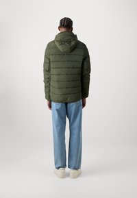 Dunkelgrüne gepolsterte Jacke mit Kapuze, mit horizontaler Steppung, von hinten gesehen, kombiniert mit hellblauen Jeans und weißen Sneakers.