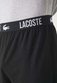 Shorts noirs avec une taille élastique portant le texte et le logo "LACOSTE" en blanc. Le matériau est lisse avec une finition légèrement mate.