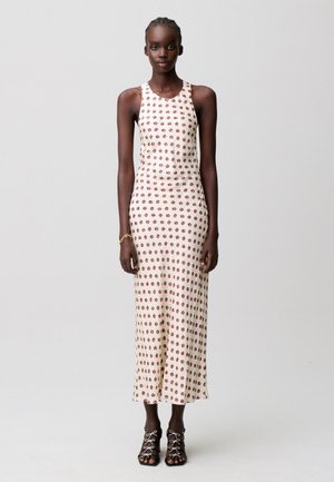 Mujer alta de piel oscura que lleva un vestido crema sin mangas hasta el tobillo con un pequeño estampado floral y sandalias negras con cordones.