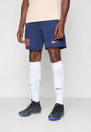 Marineblå fodboldshorts med et orange FC Barcelona-logo, parret med hvide knæhøje sokker og sorte støvler med blå accenter.