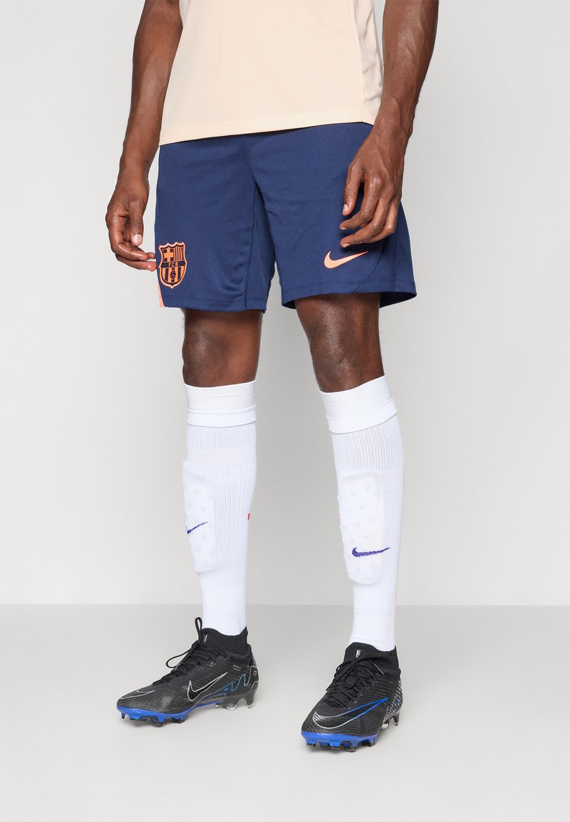 Navy soccer shorts with an orange FC Barcelona logo, paired with white knee-high socks and black cleats with blue accents.