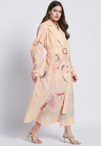 Abito trench color pesca con maniche lunghe, vita con cintura e stampe ad acquerello di libellule. Presenta bottoni e un design fluido.