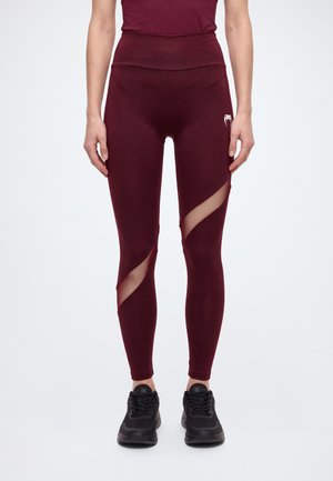 Le modèle porte un legging bordeaux taille haute avec des empiècements en mesh diagonaux et des baskets noires, sur un fond clair et uni.