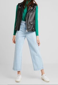 Gilet da motociclista in pelle nera sopra un dolcevita verde, abbinato a jeans a gamba larga blu chiaro e sneakers bianche. Colori vivaci e materiali classici.