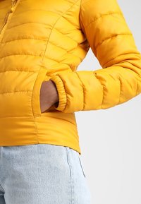 Żółta kurtka puchowa o pikowanym wzorze, z elastycznymi mankietami i bocznymi kieszeniami. Materiał wygląda na błyszczący i lekki, idealny na chłodną pogodę.