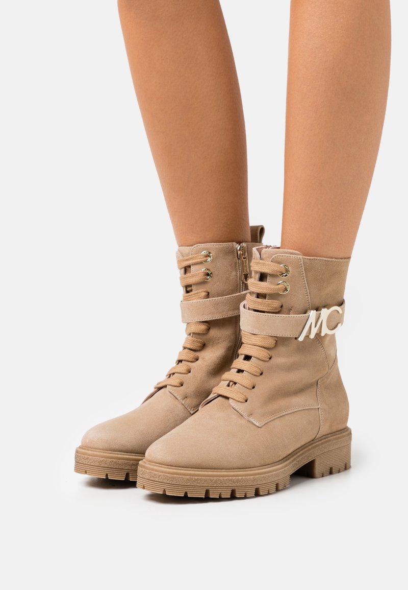 Bottines en suède beige avec une finition texturée, un laçage à l'avant, une fermeture éclair sur le côté et une sangle décorative arborant un logo "MC" doré.