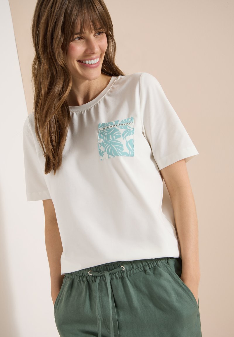 Femme souriante portant une chemise blanche à manches courtes avec une poche poitrine à motif bleu et un pantalon vert à cordon avec les mains dans les poches.