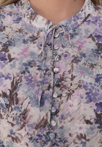 Blouse fleurie en tissu doux présentant des teintes pastel de violet, rose et bleu. Fermeture avec des boutons ronds et un détail noué à l'avant au niveau du col.