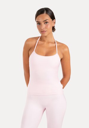 Femme aux cheveux courts et foncés, portant un haut halter rose clair et un legging moulant assorti, debout, les mains derrière le dos sur un fond uni.