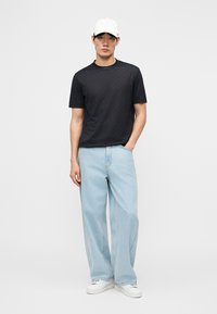 Mann mit weißer Kappe, schwarzem Kurzarmshirt, hellblauen weit geschnittenen Jeans und weißen Turnschuhen steht vor schlichtem Hintergrund.