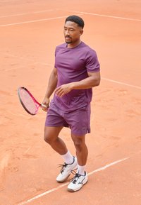 Chemise de sport violette et shorts, baskets blanches avec des accents noirs, tenant une raquette de tennis rose, debout sur un court en terre battue.
