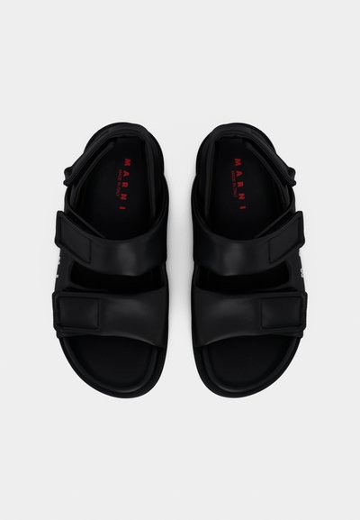 Μαύρα σανδάλια με δύο φαρδιές λουρίδες βέλκρο, κατασκευασμένα από λείο συνθετικό υλικό, με μινιμαλιστικό σχεδιασμό και ελαφρώς διαμορφωμένο πέλμα.