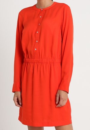 Czerwona sukienka z długim rękawem, okrągłym dekoltem, guzikami z przodu i marszczonym elastycznym pasem, noszona przez stojącą osobę.