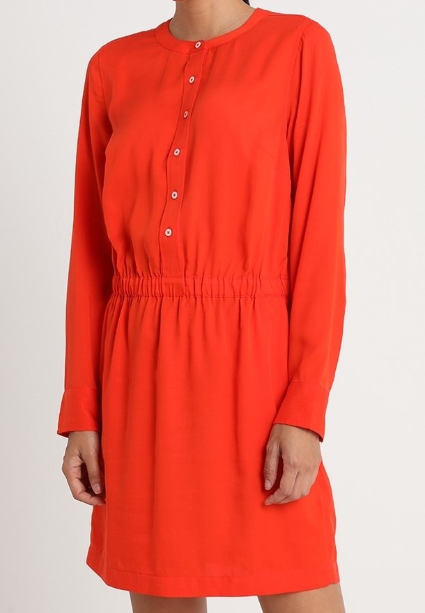 Robe rouge à manches longues avec col rond, boutons sur le devant et taille élastique froncée portée par une personne debout.