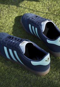 Σκούρα μπλε αθλητικά παπούτσια Adidas με ανοιχτούς μπλε ρίγες και ταμπελάκι στη φτέρνα, τοποθετημένα σε πράσινη χλοοτάπητα.