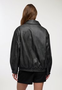 Giacca di pelle nera con una vestibilità leggermente oversize, colletto ampio, vita elastica e texture liscia. Cuciture visibili e dettagli con bottoni.