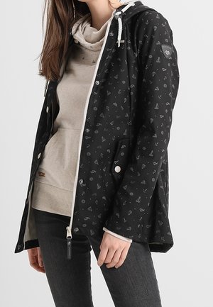 Frau trägt eine schwarze Kapuzenjacke mit weißen nautischen Mustern über einem beigen Hoodie und dunklen Jeans, steht vor einem einfarbigen Hintergrund.