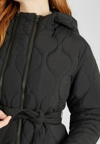 Chaqueta de plumas acolchada negra con capucha, que presenta dos cremalleras en el frente, una cintura ceñida con cinturón y un patrón de ondas texturizado.