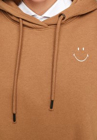 Sudadera de algodón marrón con un diseño de cara sonriente en la parte delantera, cordones ajustables y una textura suave. Presenta acentos negros en las puntas de los cordones.