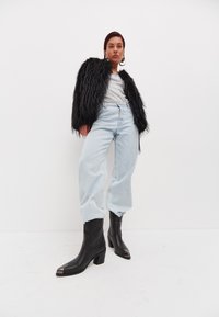 Veste en fausse fourrure noire, t-shirt gris, jean taille haute bleu clair, et bottines noires à bout carré et talon bloc.