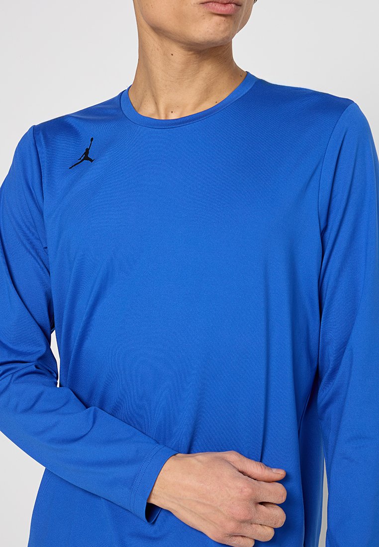 T-shirt de sport à manches longues de couleur bleu royal uni avec un col rond, arborant un logo Jumpman noir sur la poitrine gauche. Texture de tissu lisse.