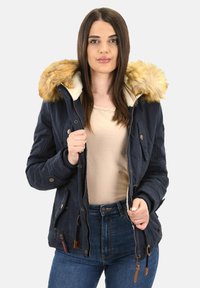 Vrouw in een navyblauwe winterjas met een bontafgewerkte capuchon, een beige bovenstuk en blauwe jeans, die de jas gedeeltelijk open heeft ritsen.