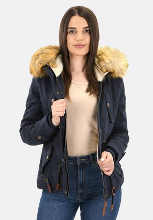 Vrouw in een navyblauwe winterjas met een bontafgewerkte capuchon, een beige bovenstuk en blauwe jeans, die de jas gedeeltelijk open heeft ritsen.