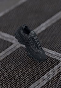 Black running shoe with mesh and synthetic upper, featuring a rounded toe, textured accents, and a cushioned sole. Brand logo visible on side.