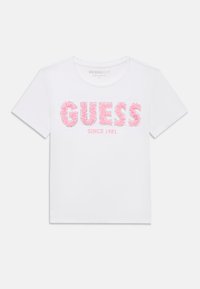 T-shirt blanc à manches courtes avec "GUESS" en lettres florales roses et "SINCE 1981" imprimé en dessous en texte rose.
