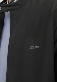 Simább szövetből készült fekete dzseki, magas gallérral, patentos záródással és az oldalon elhelyezett ezüst színű "JACK & JONES" logós címkével.