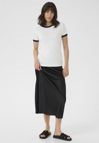 T-shirt blanc à manches courtes avec des bordures noires, associé à une jupe maxi noire fluide. Des sandales à glissière noires complètent la tenue.