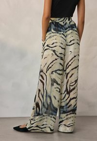 Pantalons larges légers et transparents en beige avec un motif abstrait de rayures de tigre noires et bleues. Dotés d'une taille froncée et de poches latérales.