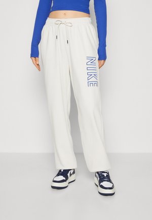 Osoba ubrana w białe spodnie dresowe Nike z niebieskim logo na udzie, niebieski krótki top z długim rękawem oraz białe i granatowe sneakersy stojąca na szarej podłodze.