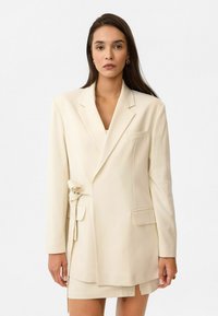 Beige op maat gemaakte blazer met een ceintuur in de taille, notch revers en een enkele borstzak. Glad materiaal, rechte pasvorm en lange mouwen.