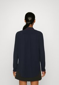 Marineblaues Langarmshirt mit abgerundetem Saum, Knopfmanschetten und kontrastierenden roten Nähten am Kragen. Glattes, leichtes Material.