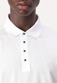 Witte poloshirt met een geribde kraag, vijf zwarte knopen en subtiele logo- borduursel op de linker borst. Soepele katoenen textuur.